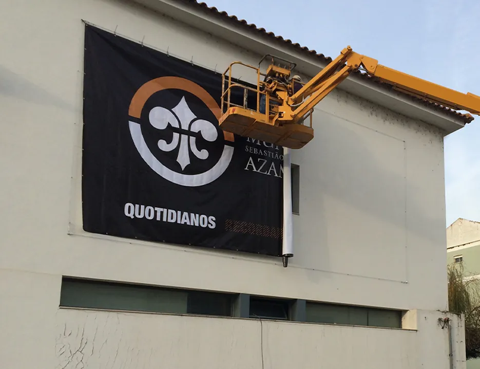 Lona com o logótipo Quotidianos numa fachada, com uma plataforma elevatória ao lado